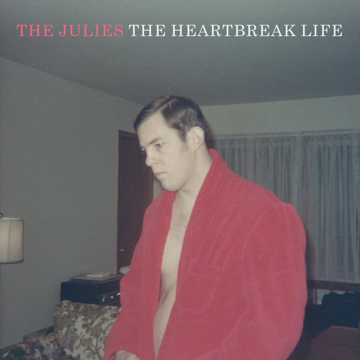 Album cover for The Heartbreak Life by The Julies, showing a man in a red bathrobe standing in a living room.