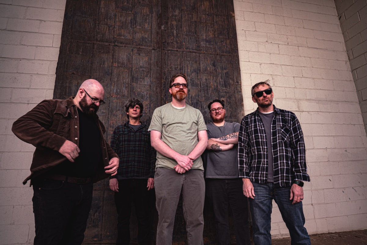Five men standing against a brick wall, with one man in the foreground wearing glasses.