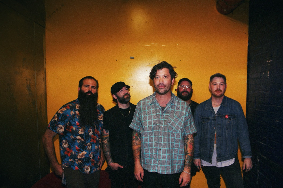 Five men standing against a yellow wall with tattoos visible on their arms.