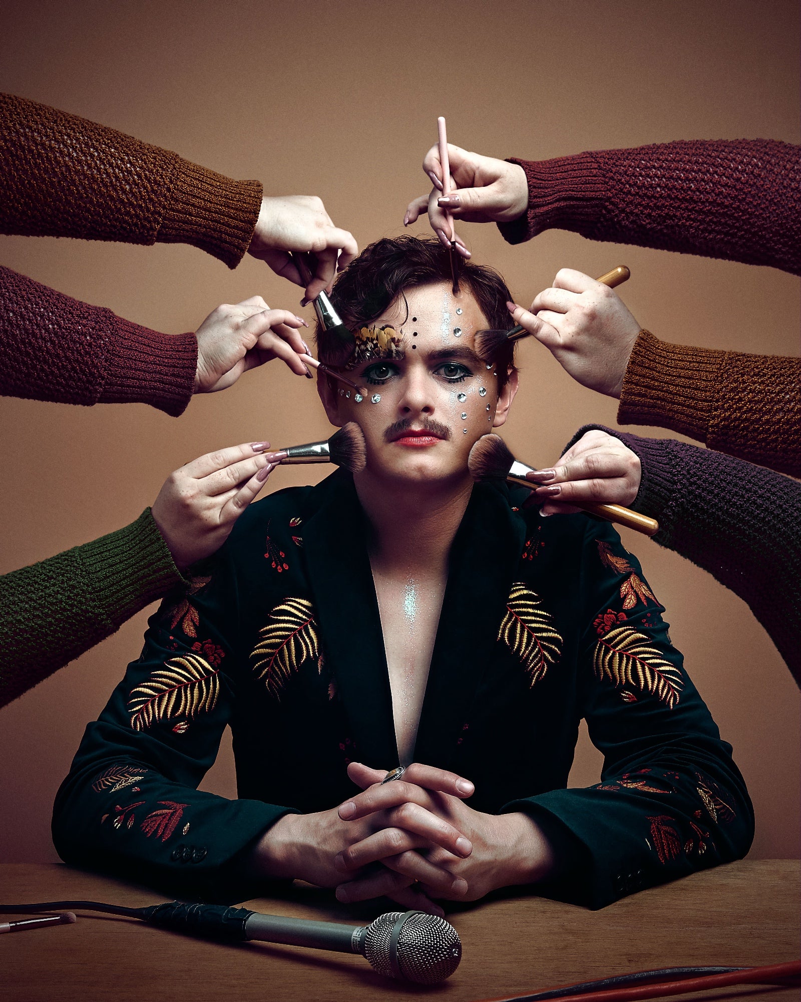 Person in a patterned jacket sitting at a table with makeup brushes applied to their face by multiple hands, microphone in front.
