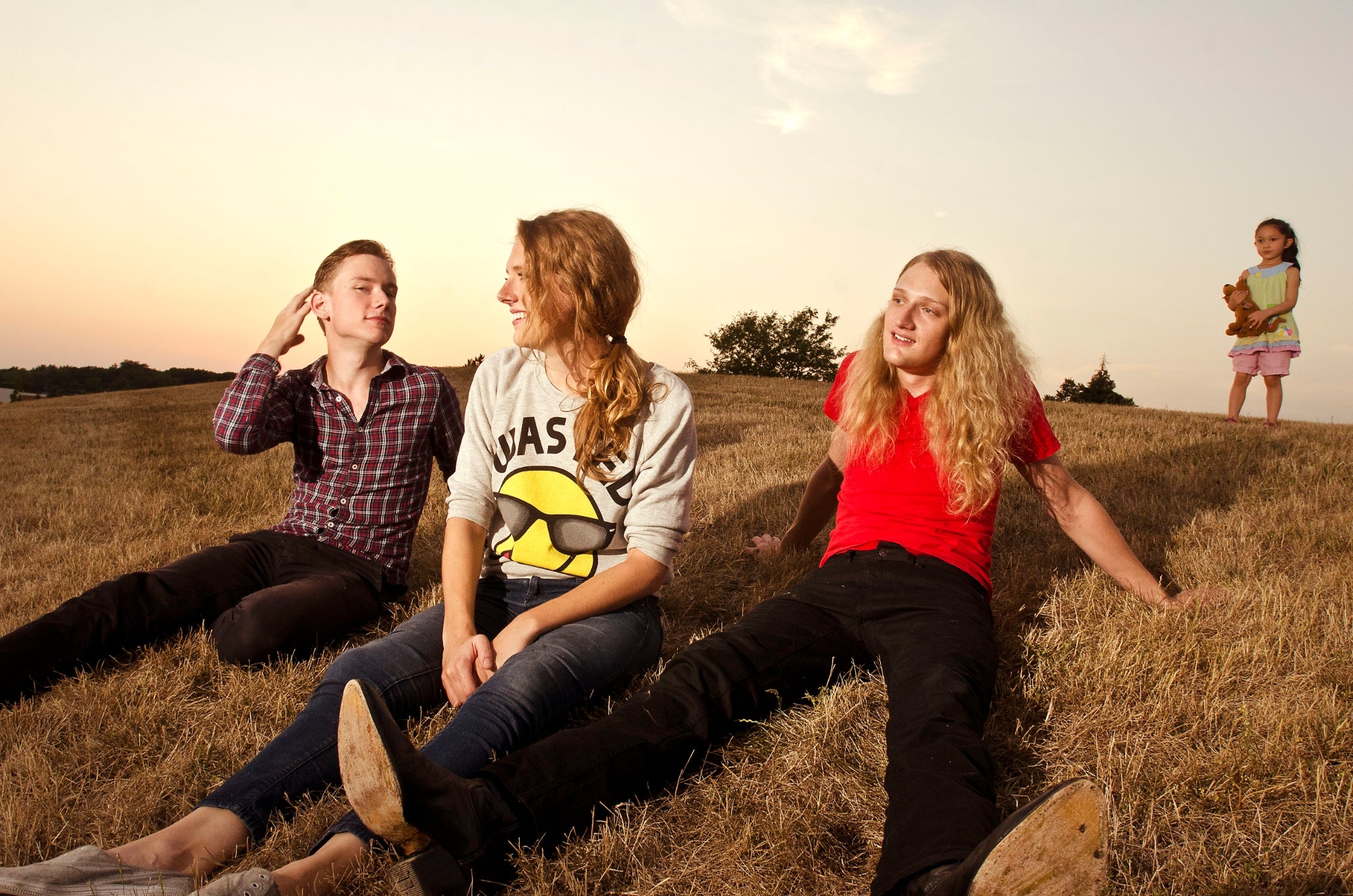 Three people sitting on a grassy hillside at sunset with a child holding a teddy bear in the background.